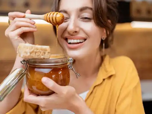 Smiling woman holding a jar of honey with a honey dipper, enjoying fresh honeycomb.